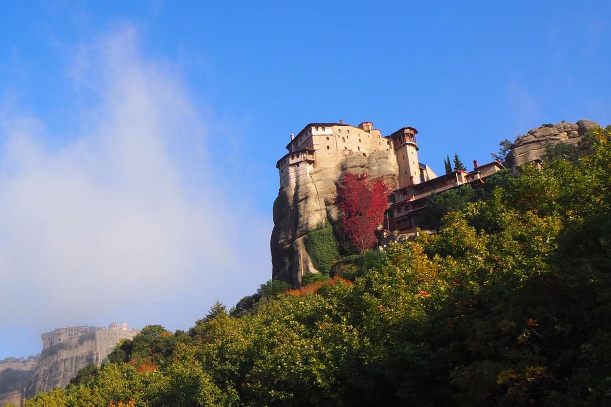 Greece tours: a Meteora monastery perched on a tall rock above green forest under a blue sky, with another monastery in the mist far behind.